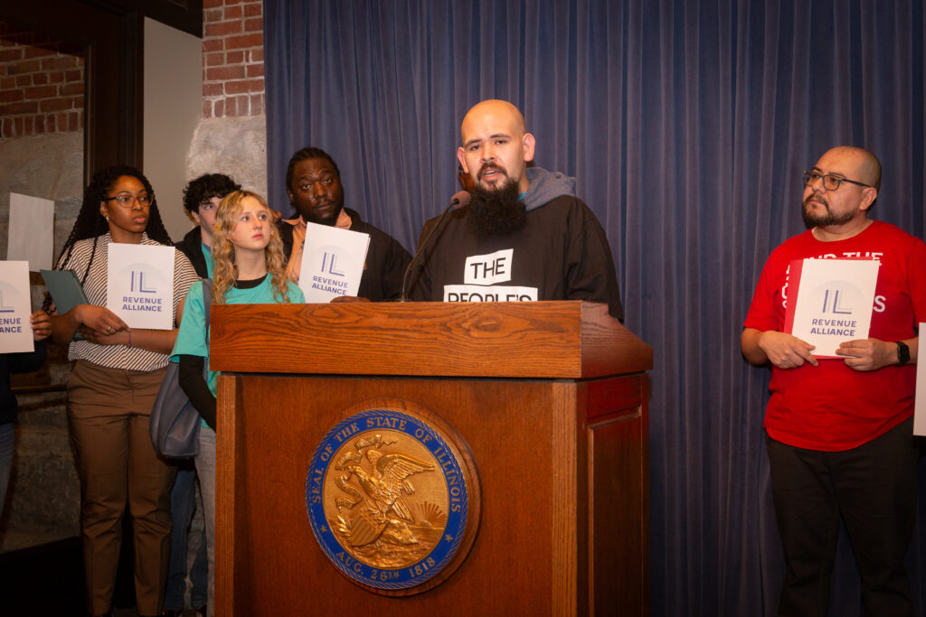 Bruno Garcia, a member of The People's Lobby, stands at a podium talking. He is surrounded by other speakers holding Illinois Revenue Alliance posters.