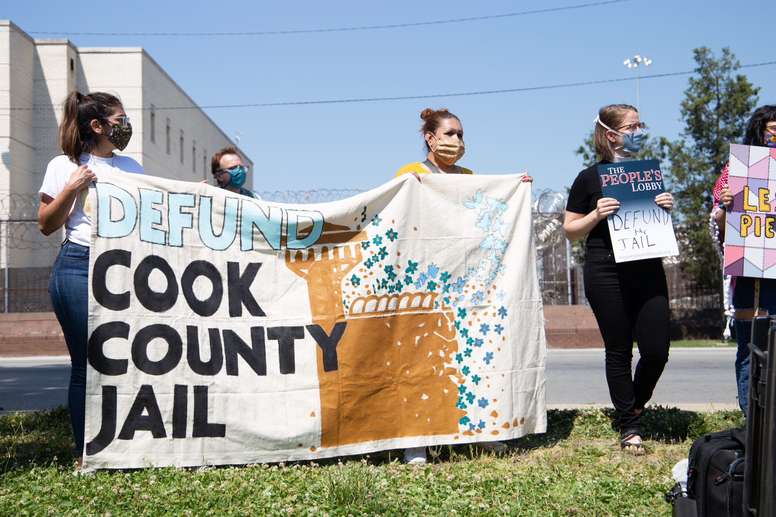 A few people wearing masks on the side of an empty road holding a cloth sign that reads “Defund Cook County Jail”.