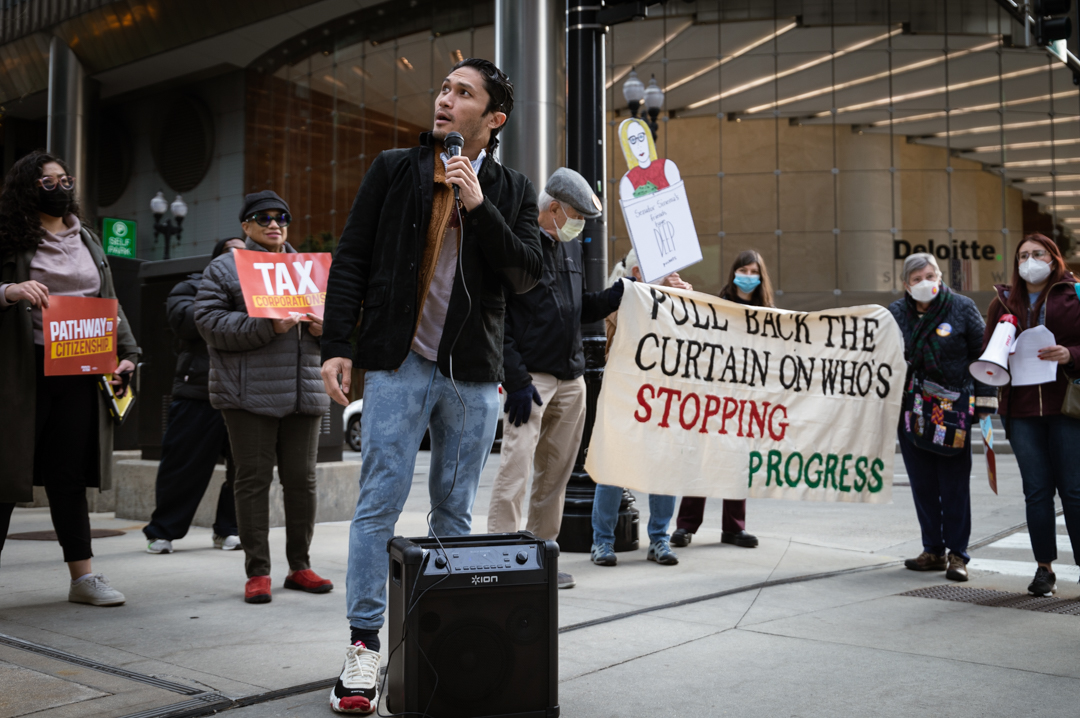 A person holding a mic connected to an amplifier looking upwards while surrounded by a few activists holding signs.