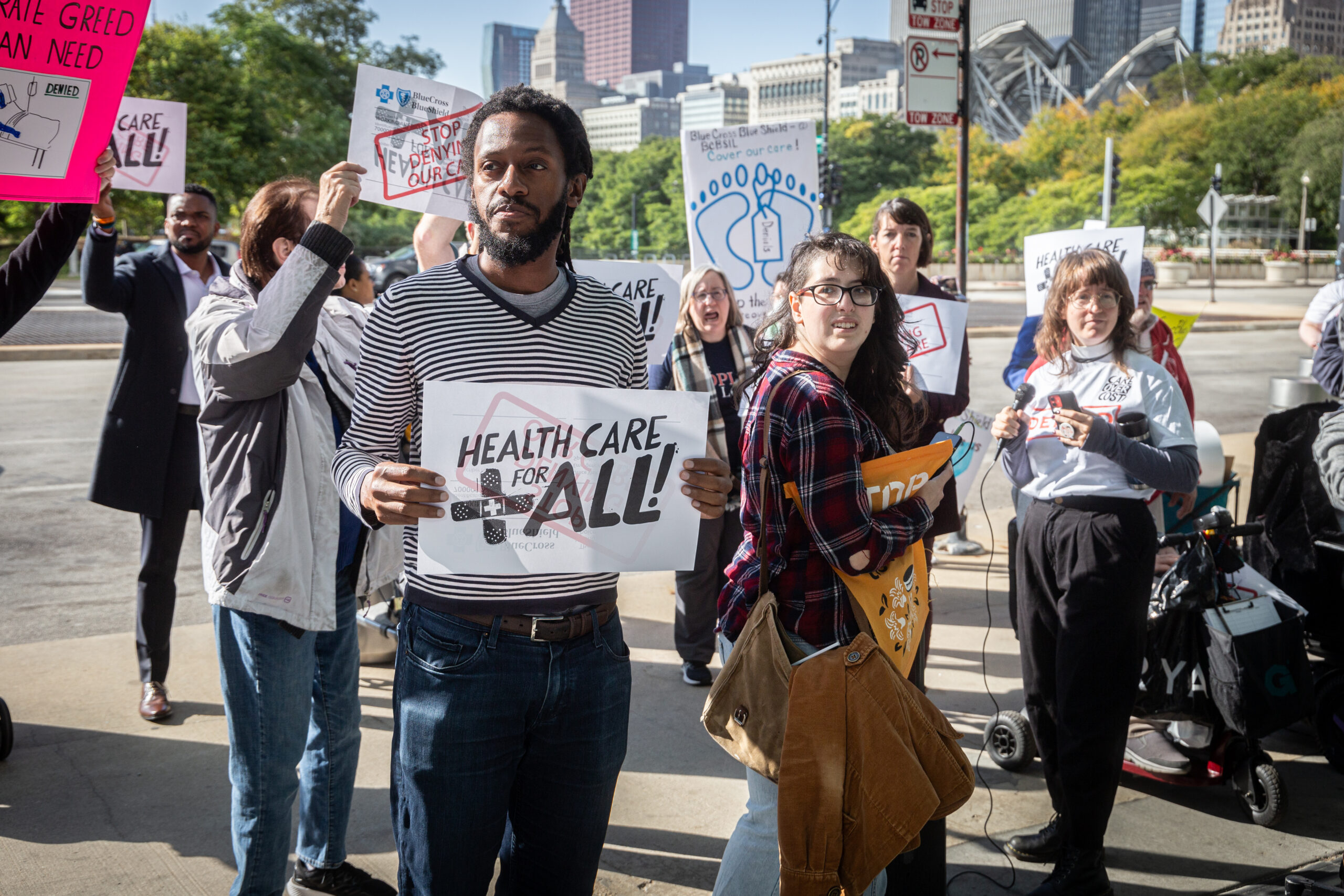 A group of people outdoors holding that read “Healthcare for all” looking in different directions.