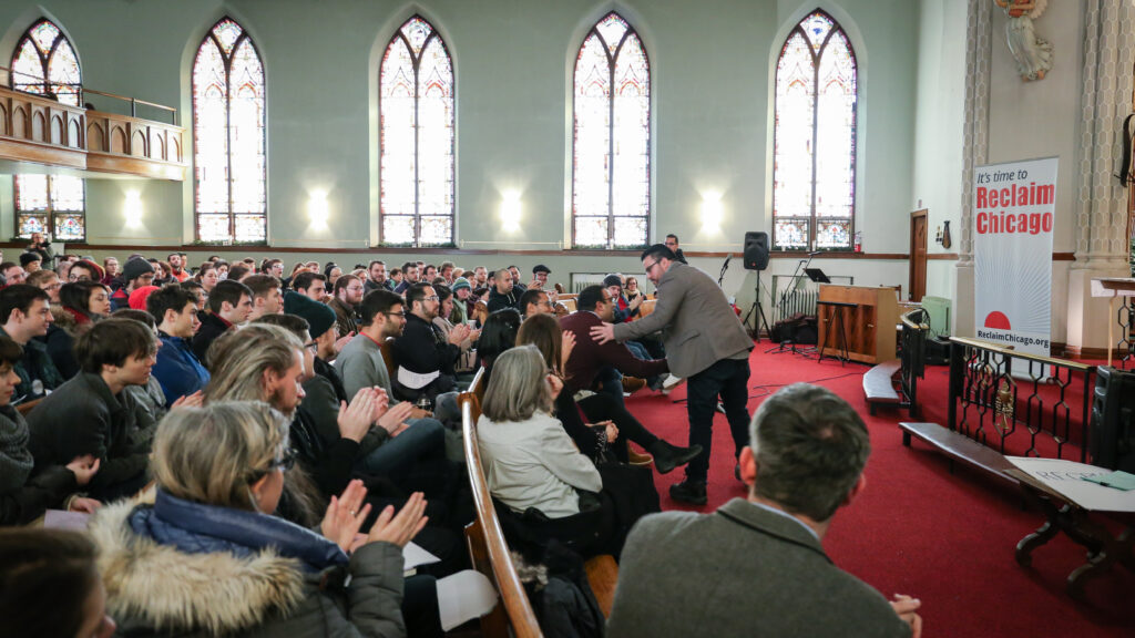 A standing person shaking hands with a seated person surrounded by a large group of people seated in a church clapping their hands.