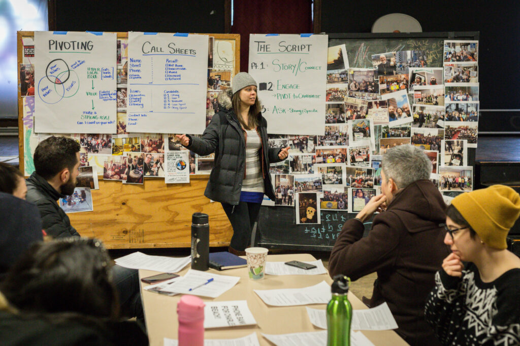 A person in front of a board with several photos conducting an informational session for people seated around a table.