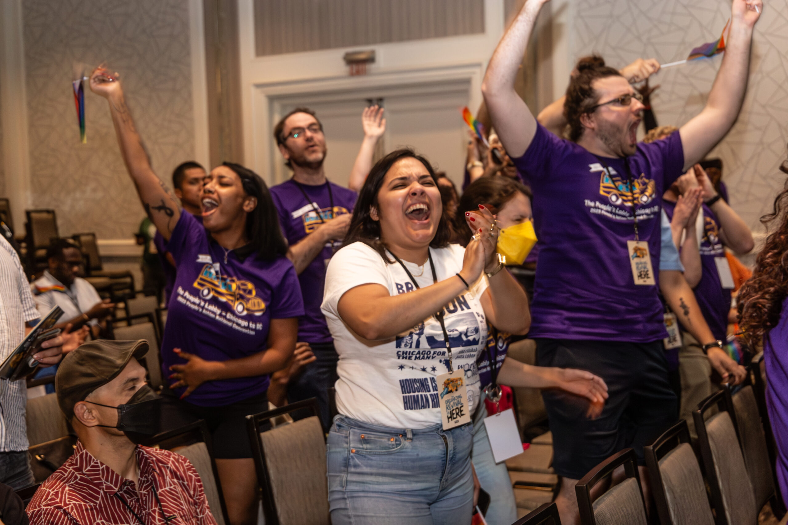 A group of people in an event hall celebrating and cheering with some holding pride flags.