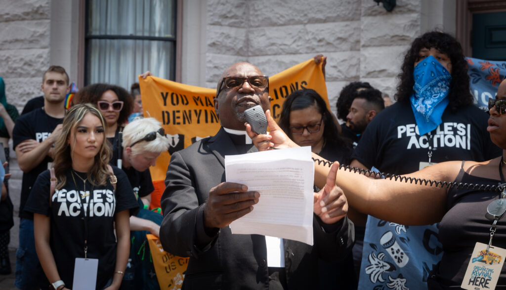 A person holding a few papers and speaking into a mic surrounded by other activists.