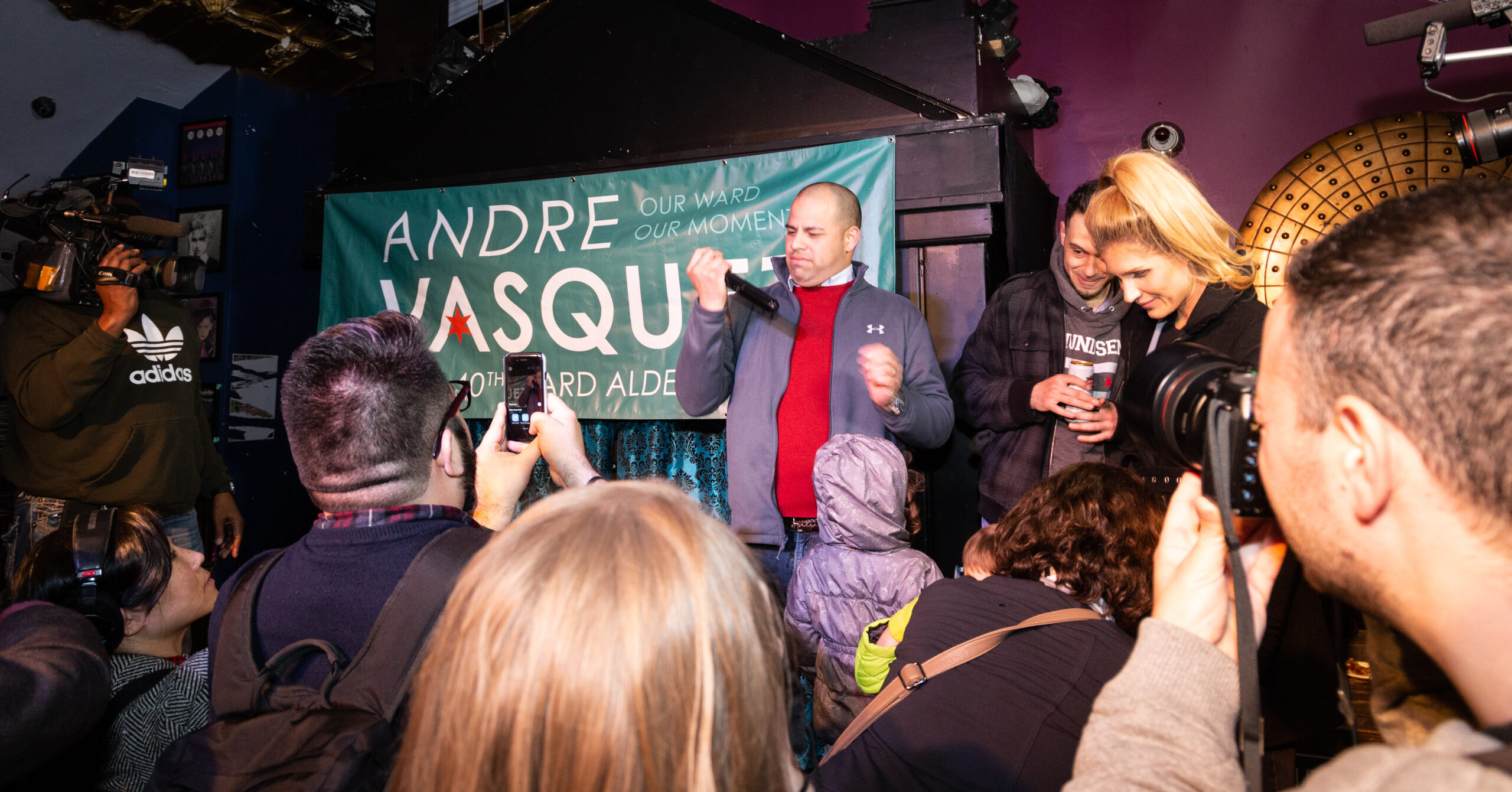 Person on a stage with a mic surrounded by a few audience members, some holding cameras.