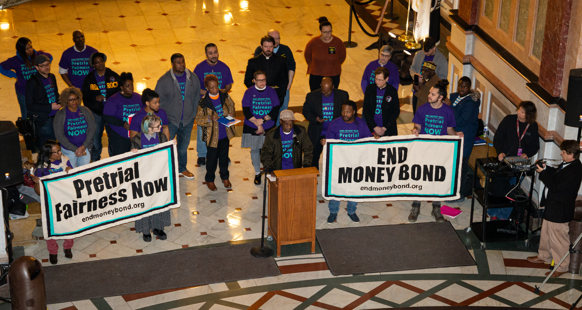 Aerial photograph of a person in front of a podium surrounded by several people wearing “Pretrial Fairness Now” tshirts and holding two large signs.