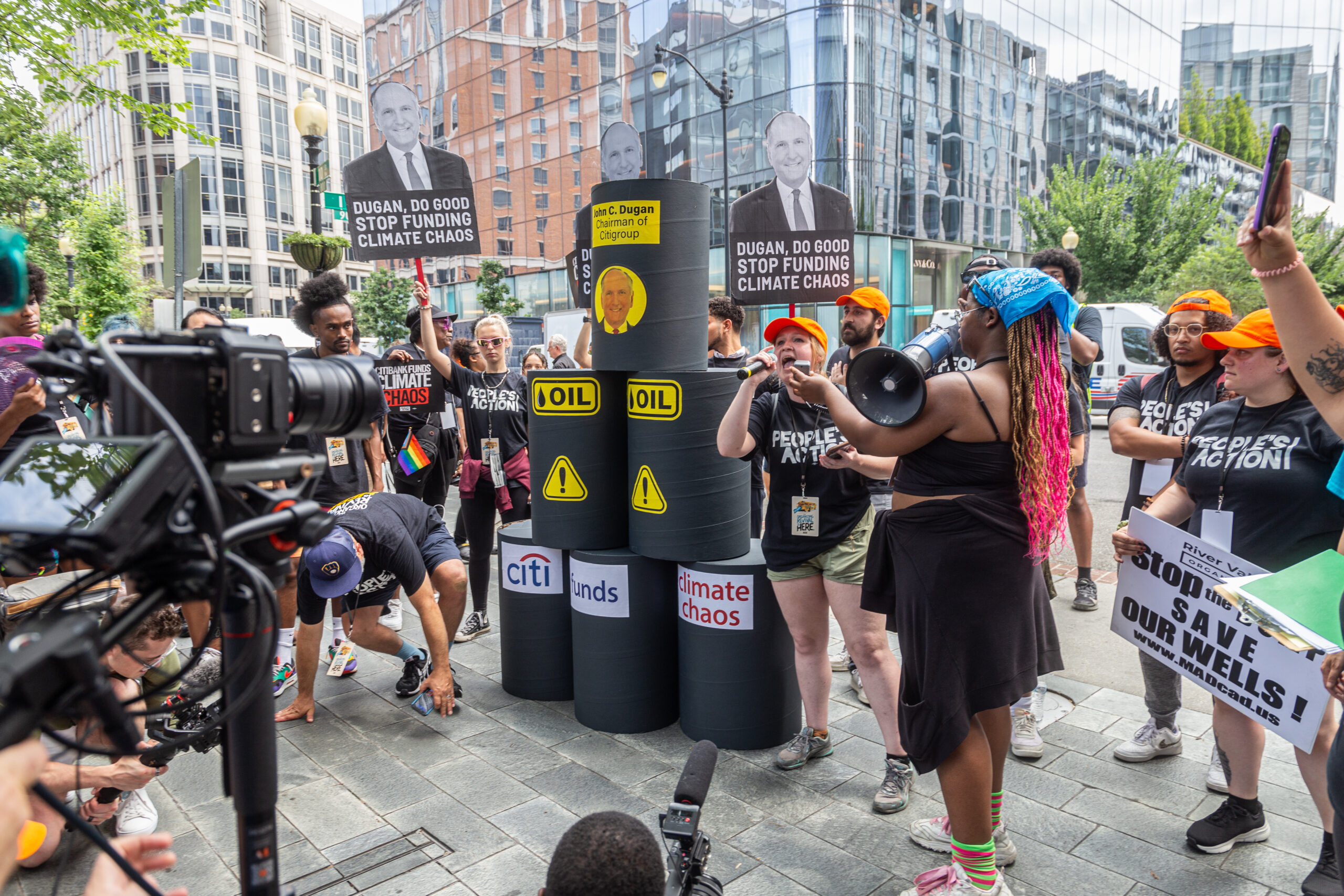 Group of climate activists in a downtown area holding signs in front of six large oil cans with text that reads “Citi funds climate chaos”.