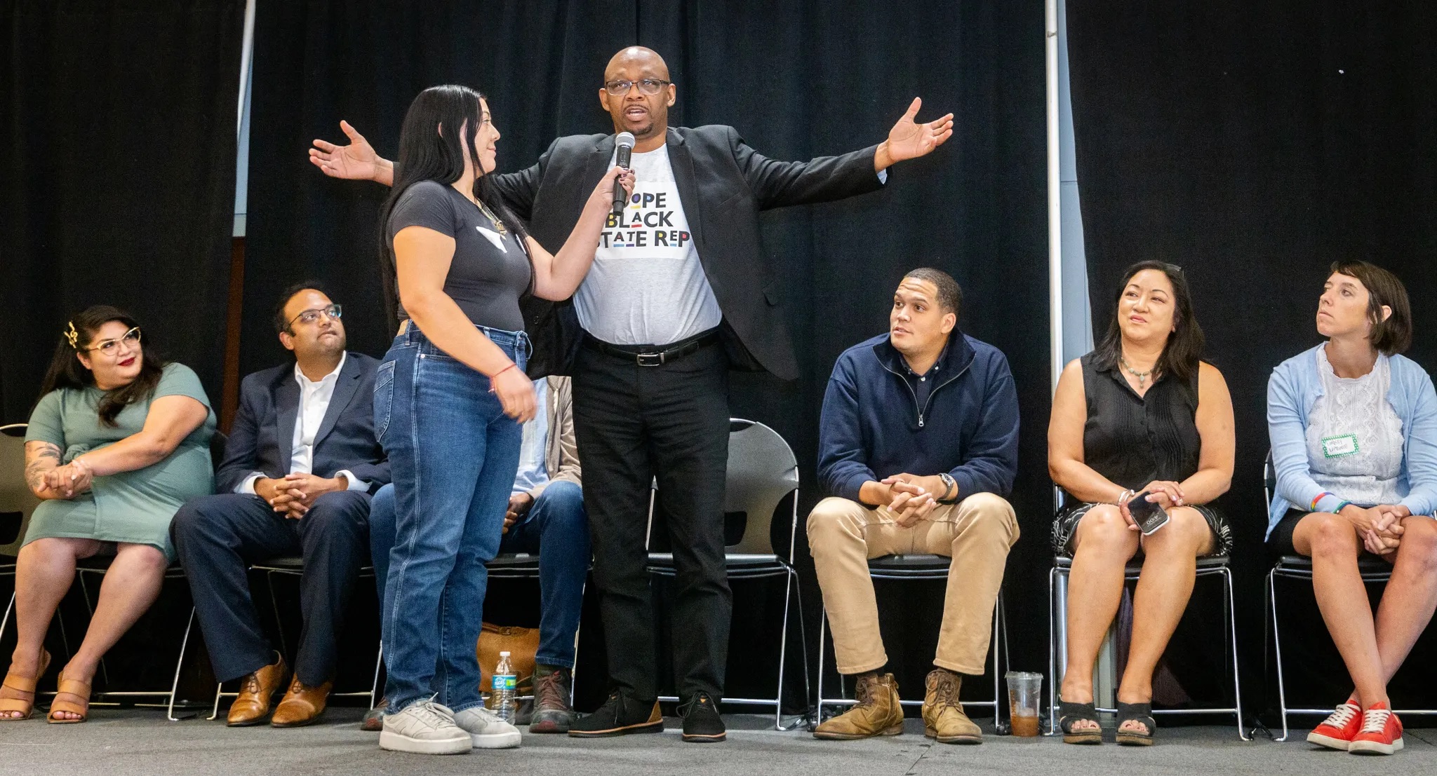 Person on a stage with arms outstretched surrounded by others on chairs looking at them.