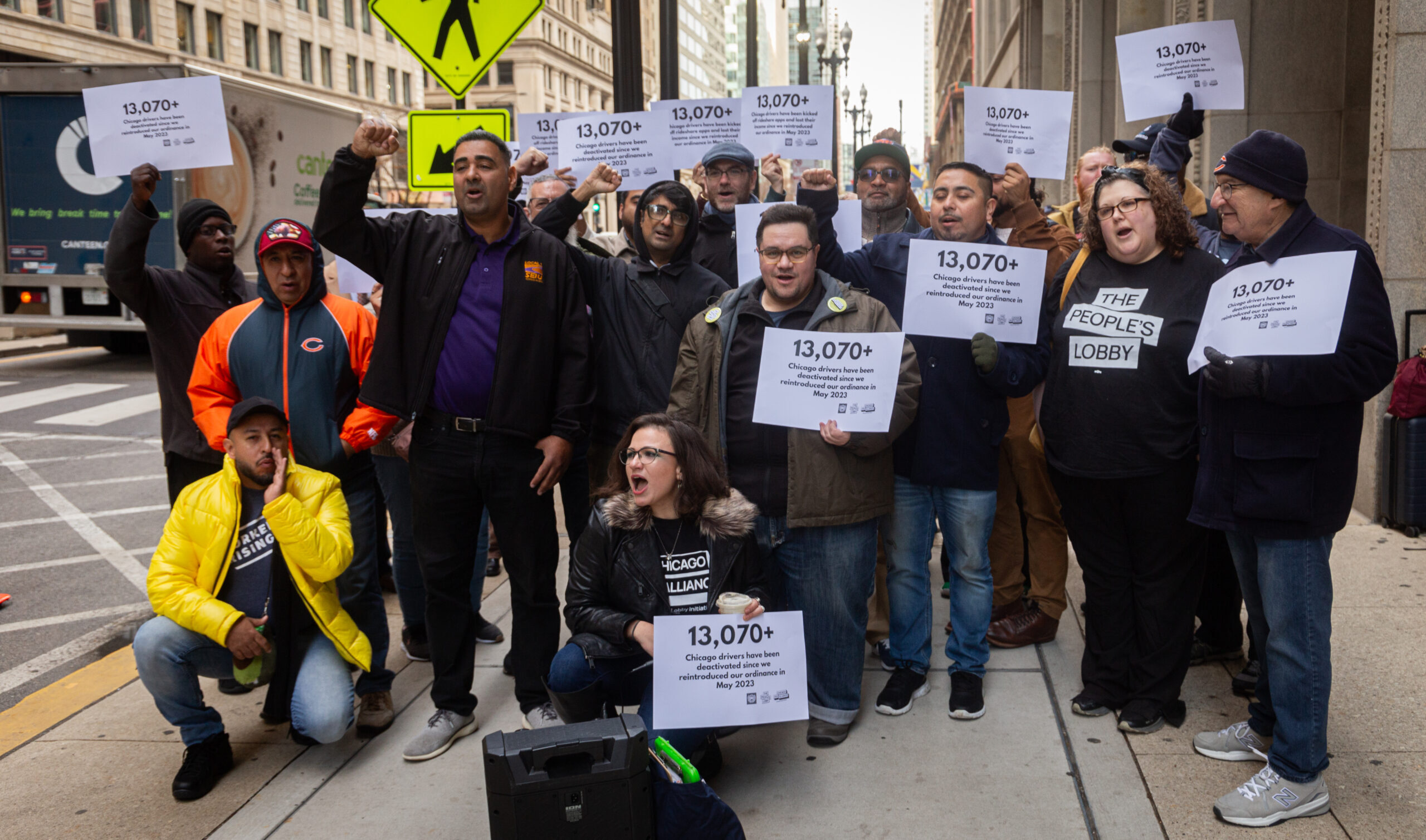 Rideshare workers protesting outside city hall, carrying signs that say 13,000+ drivers have been deactivated since May 2023.