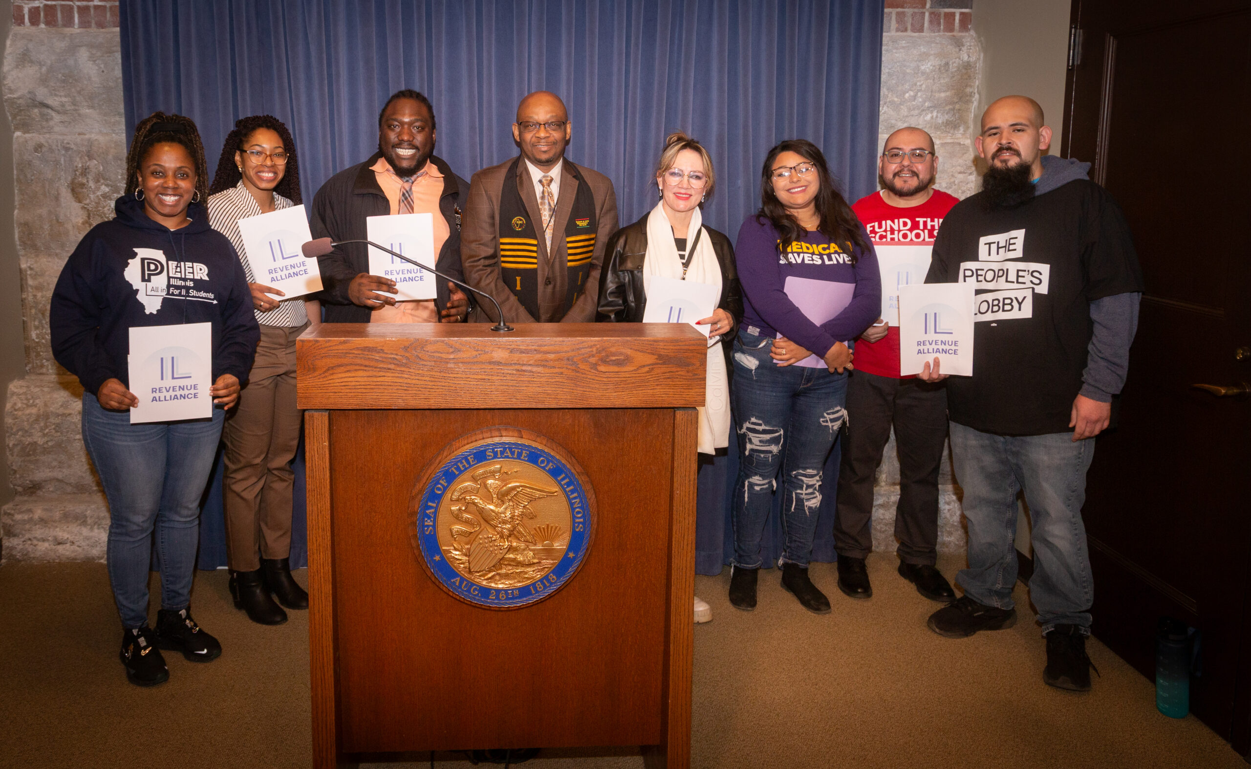 Speakers with the Illinois Revenue Alliance stand around a podium with a blue background behind them. They are holding posters that say Illinois Revenue Alliance.