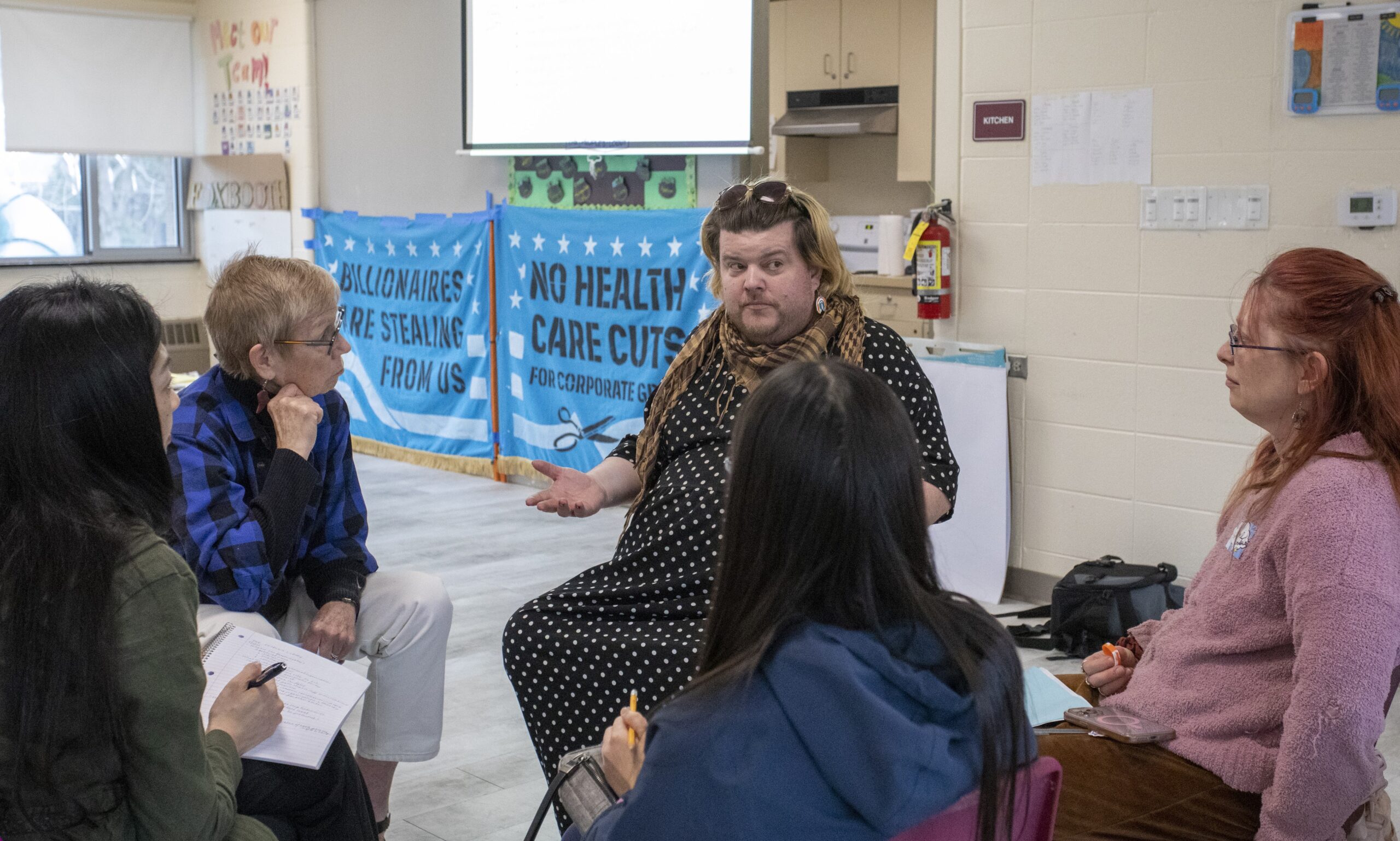 Five people sit in chairs in a circle, discussing. Two blue banners behind them say "Billionaires Are Stealing From Us" and "No Health Care Cuts."
