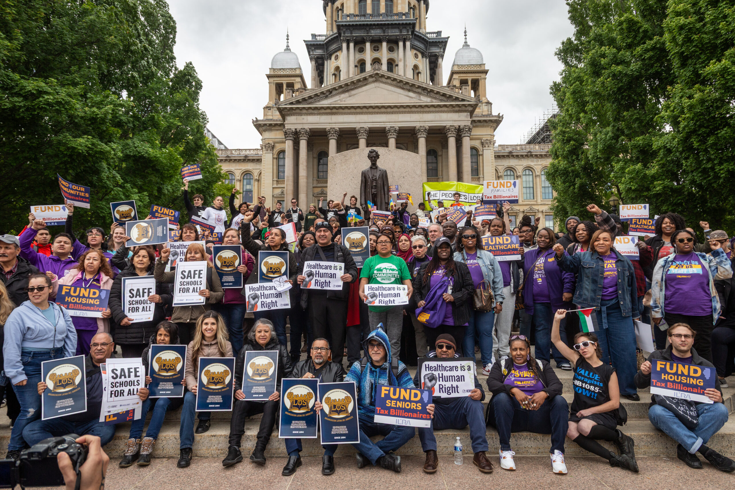 A crowd of 250+ people stand outside the Illinois State Capitol building with signs that say "Fund Our Communities."